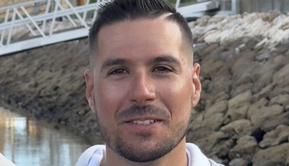 Man in gray hoodie smiling, with palm trees and buildings in the background.